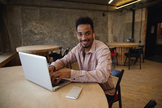Cheerful young bearded dark skinned male in casual clothes sitting over coworking space, looking at camera with wide charming smile and keeping hands on keybord of his modern laptop