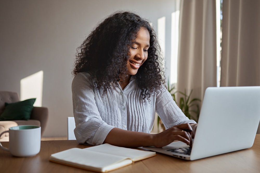 Attractive positive young Afro American woman freelancer working remotely, keyboarding on generic laptop, sitting at home with copybook and mug on table, typing electronic message online, smiling