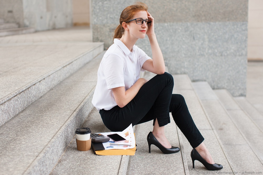 Portrait of pensive young Caucasian businesswoman wearing glasses sitting on staircase with papers, calculator and coffee cup. Work life balance concept