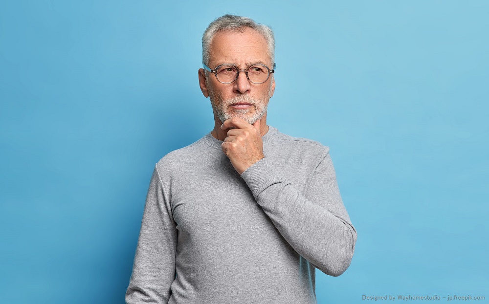 Old age and thoughts concept. Serious grey haired man holds chin and concentrated with pensive expression somewhere dressed in casual turtleneck isolated over blue background thinks over problems