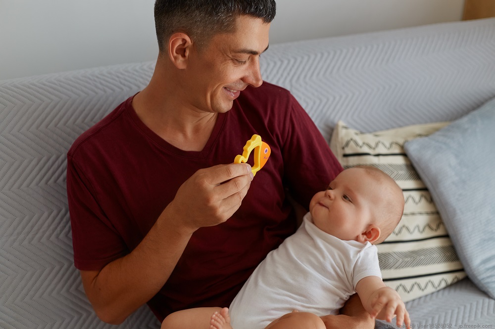 Happy father holding toy and playing with his baby boy or girl while sitting on sofa, smiling man wearing maroon t shirt showing to child orange fish, happy parenthood.